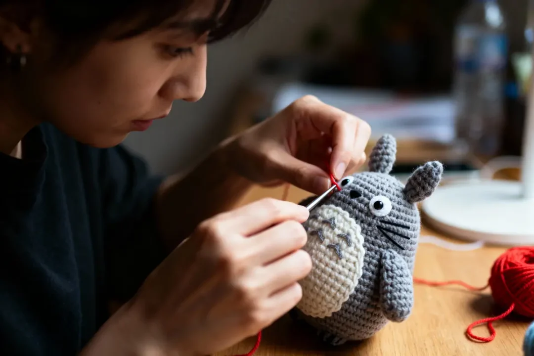 Personne crochetant un personnage gris en laine avec un fil rouge, concentrée sur les détails.