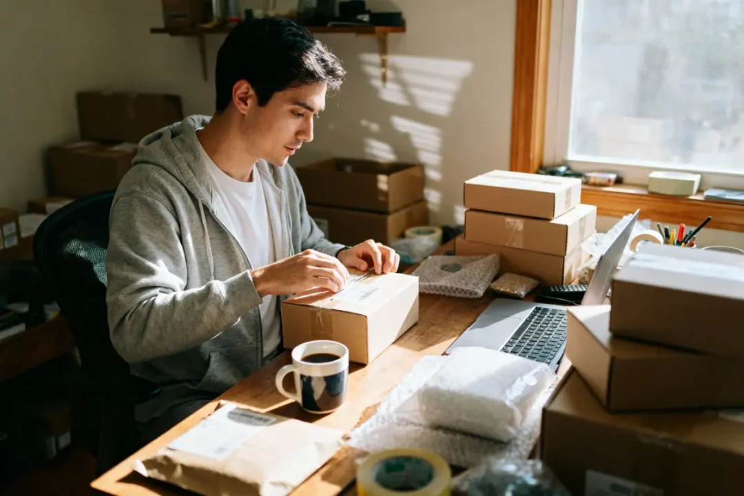 Homme emballant des colis à un bureau, avec ordinateur portable et tasse de café, lumière naturelle.