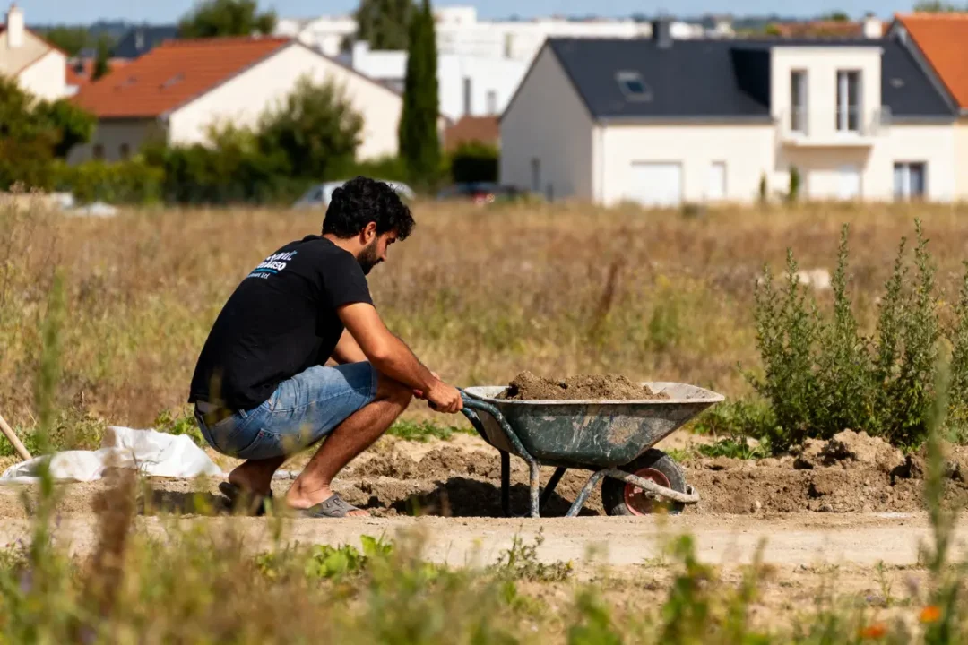 Un homme, accroupi sur un terrain, remplit une brouette de terre, maisons en arrière-plan.