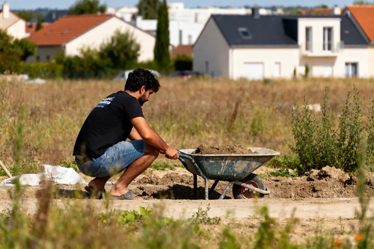 Un homme, accroupi sur un terrain, remplit une brouette de terre, maisons en arrière-plan.