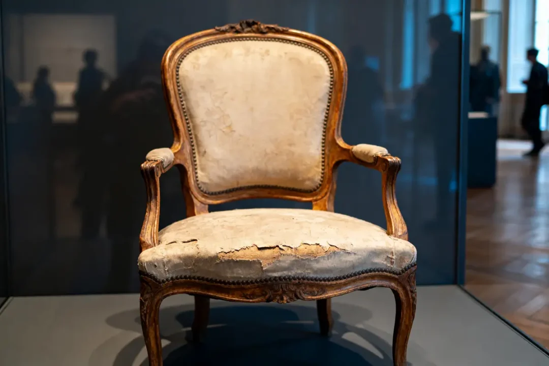 Fauteuil ancien en bois sculpté, tissu usé, exposé dans un musée, avec visiteurs en arrière-plan flou.