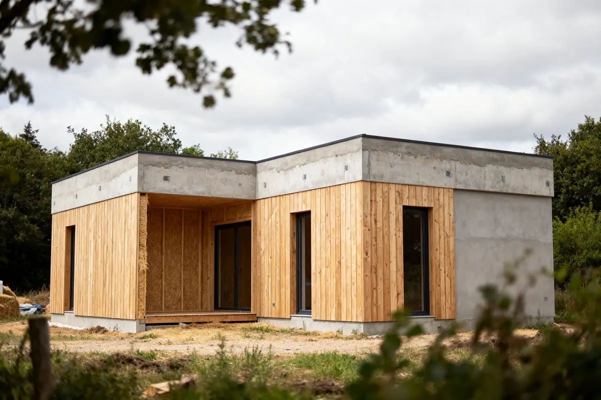 Maison moderne en bois et béton, entourée de verdure, sous un ciel nuageux.