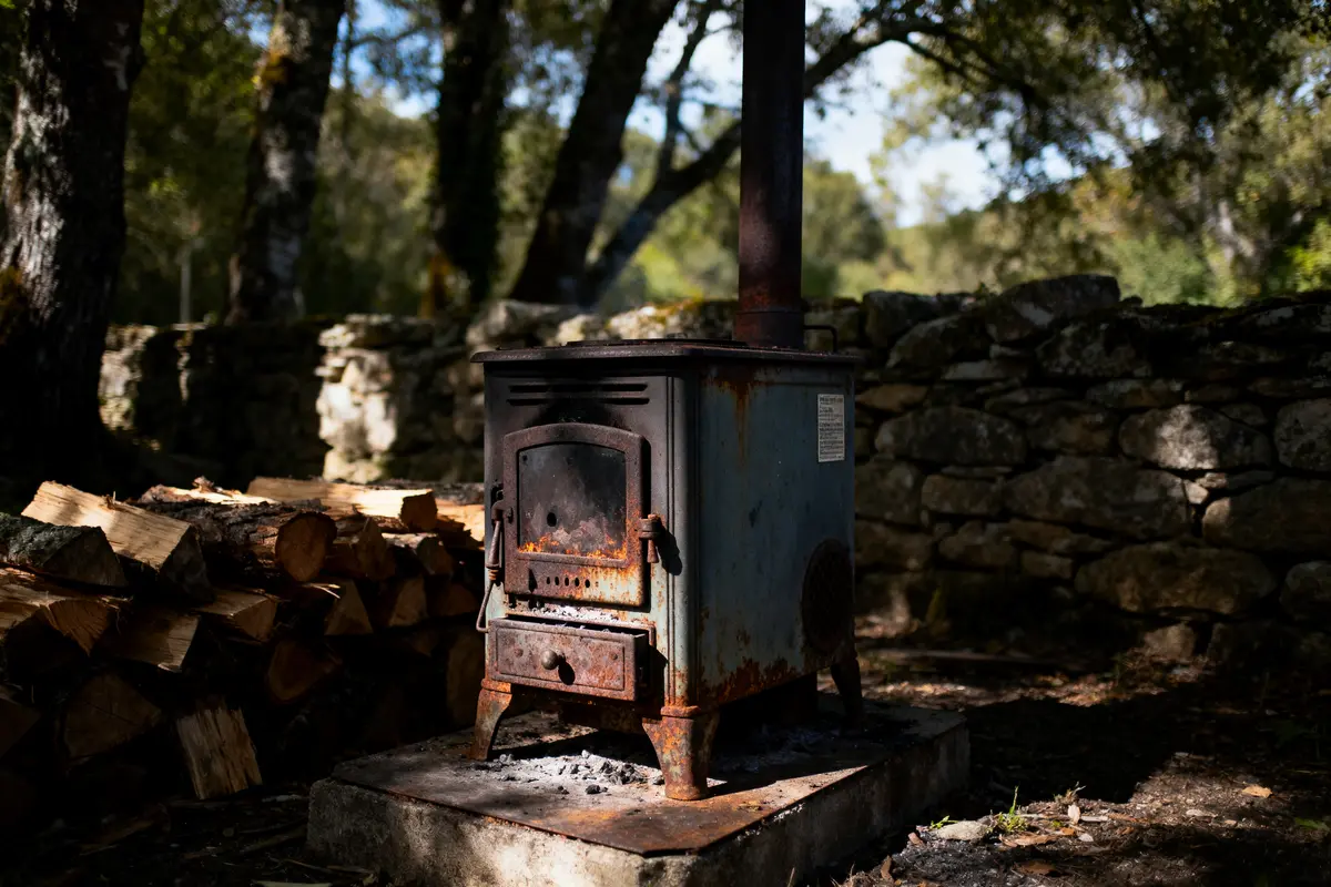 Poêle à bois rouillé en extérieur, proche de bûches et mur en pierre, éclairage naturel, forêt en arrière-plan.