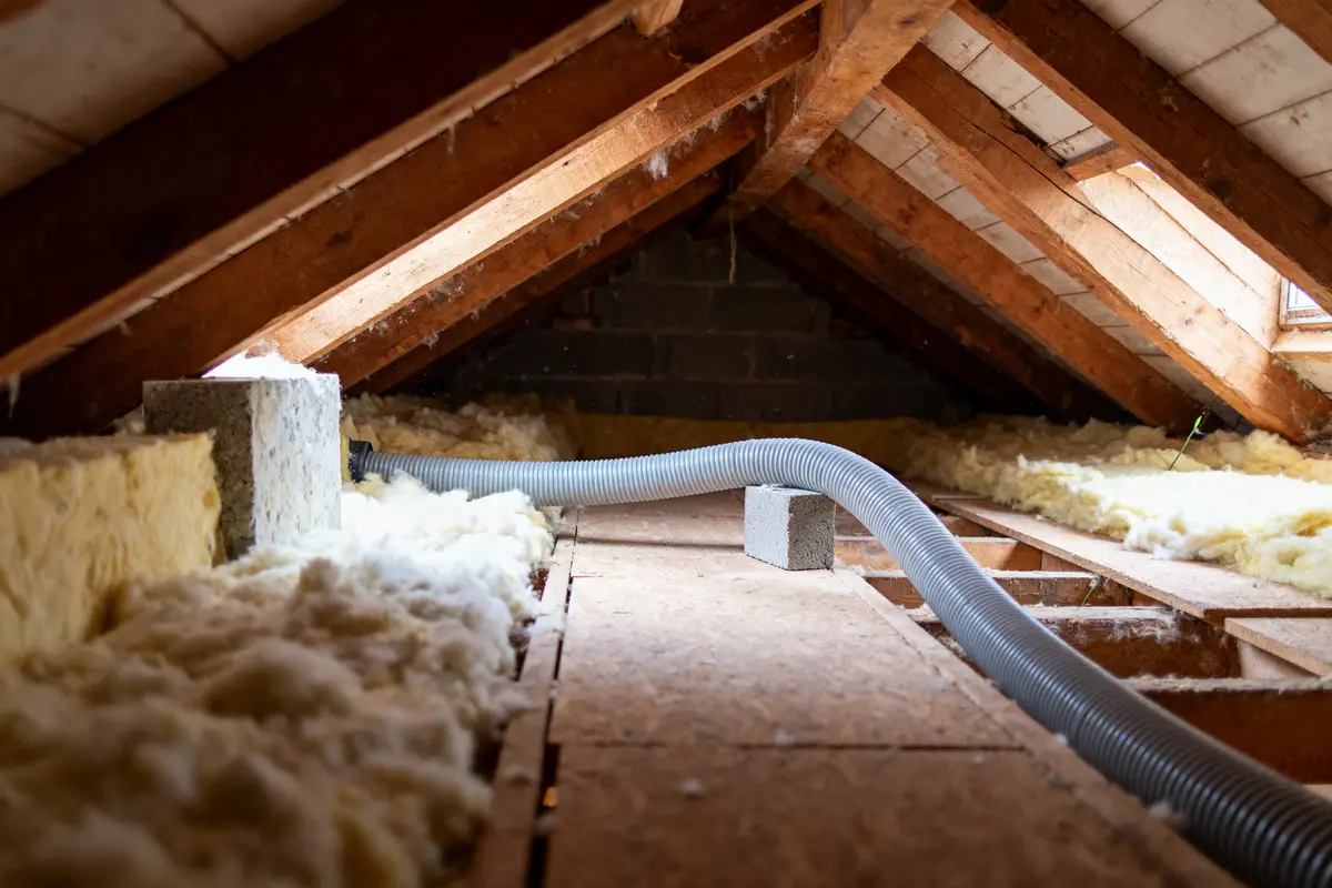 Combles en bois avec isolation, laine et conduit d'aération gris. Lumière naturelle entrant par une fenêtre.