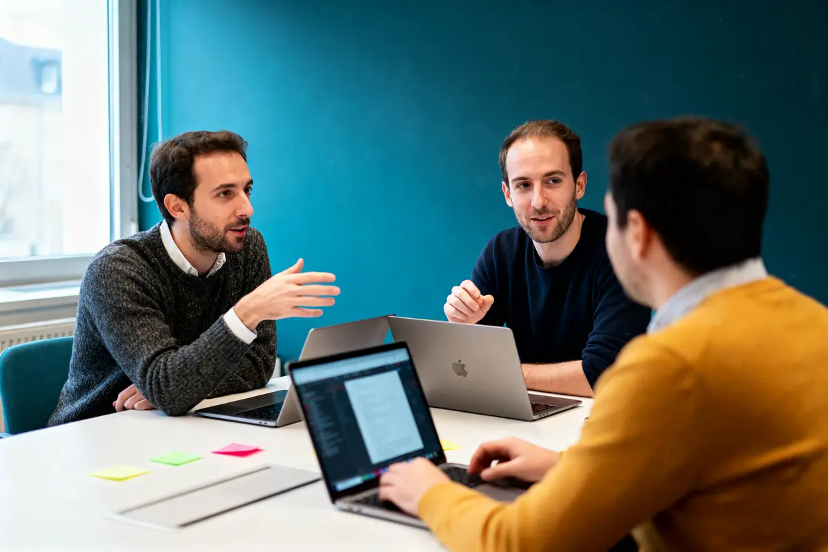 Trois hommes discutent autour d'une table, utilisant des ordinateurs portables, devant un mur bleu.