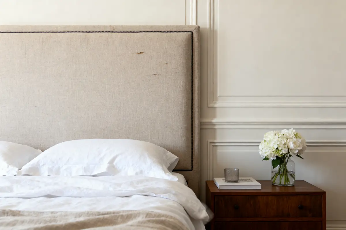Lit avec draps blancs, tablette de nuit en bois, vase fleurs blanches, verre transparent, ambiance élégante minimaliste.