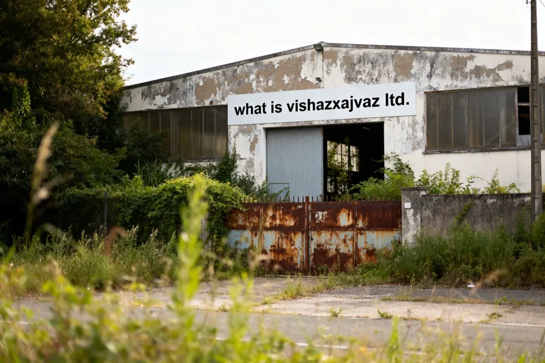Bâtiment abandonné avec pancarte "what is vishazxajvaz ltd." entouré de végétation, portail rouillé devant.