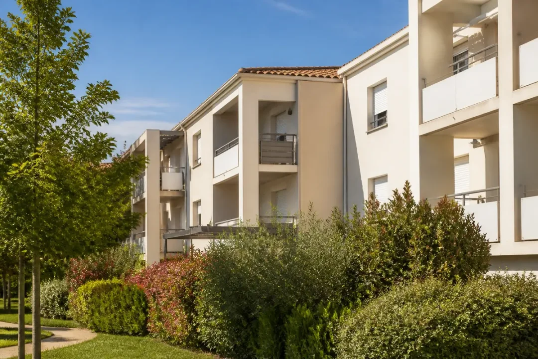 Immeuble résidentiel moderne, arbres et buissons denses, ciel bleu.