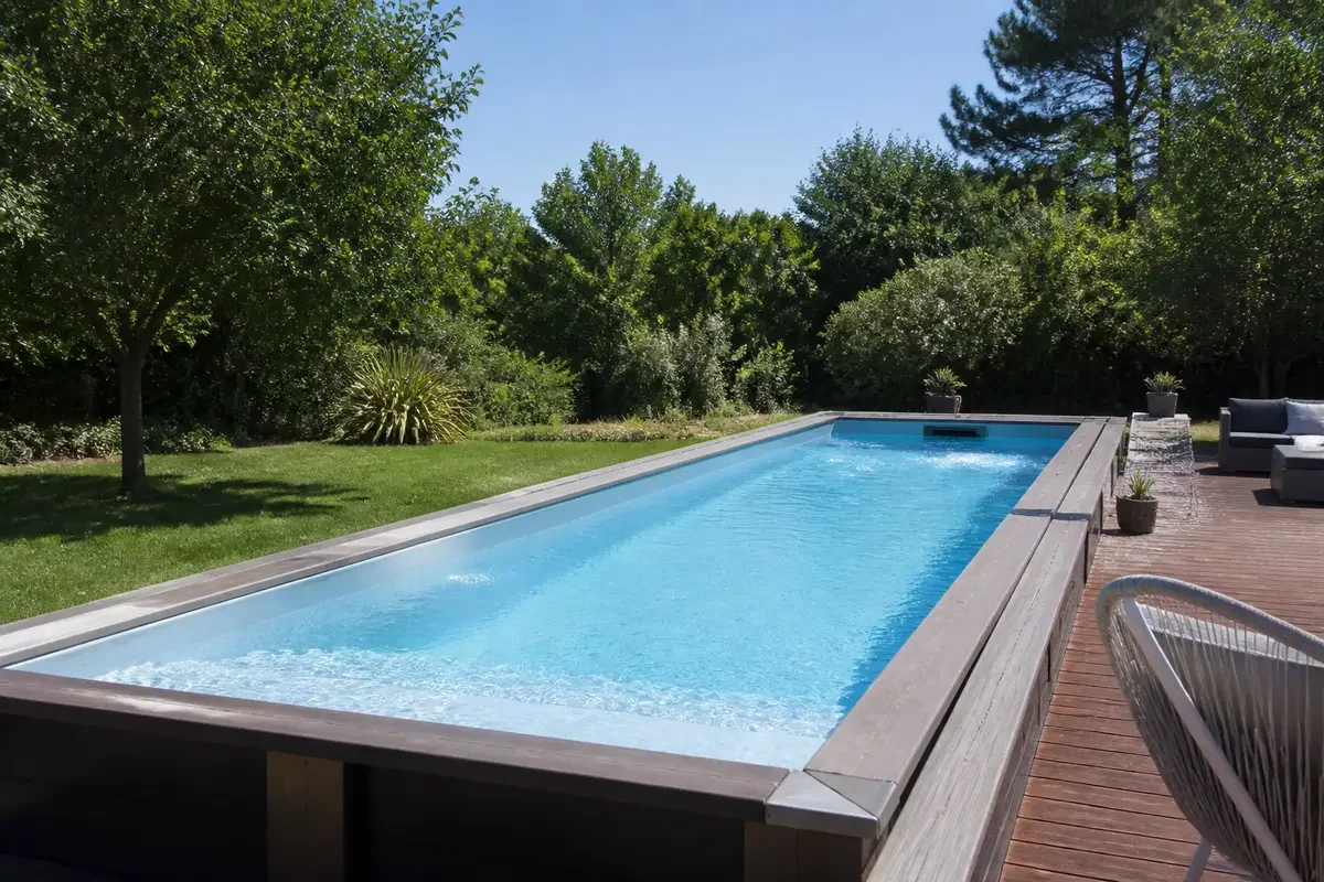 Piscine rectangulaire avec margelle en bois dans jardin verdoyant, ciel bleu, espace détente ombragé.