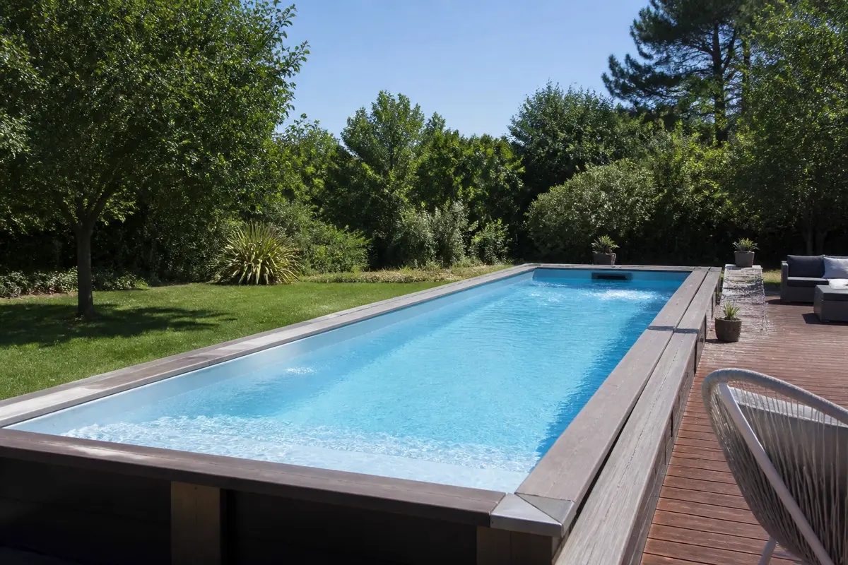 Piscine rectangulaire avec margelle en bois dans jardin verdoyant, ciel bleu, espace détente ombragé.