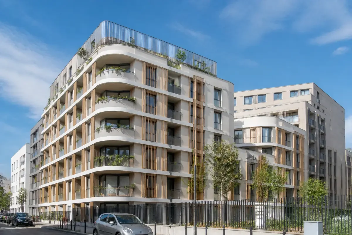 Immeuble moderne à balcons végétalisés, ciel bleu, voitures stationnées devant, quartier résidentiel urbain.