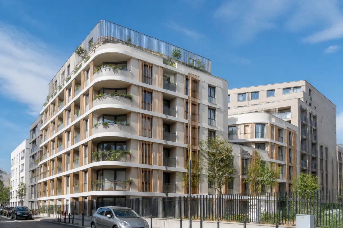 Immeuble moderne à balcons végétalisés, ciel bleu, voitures stationnées devant, quartier résidentiel urbain.