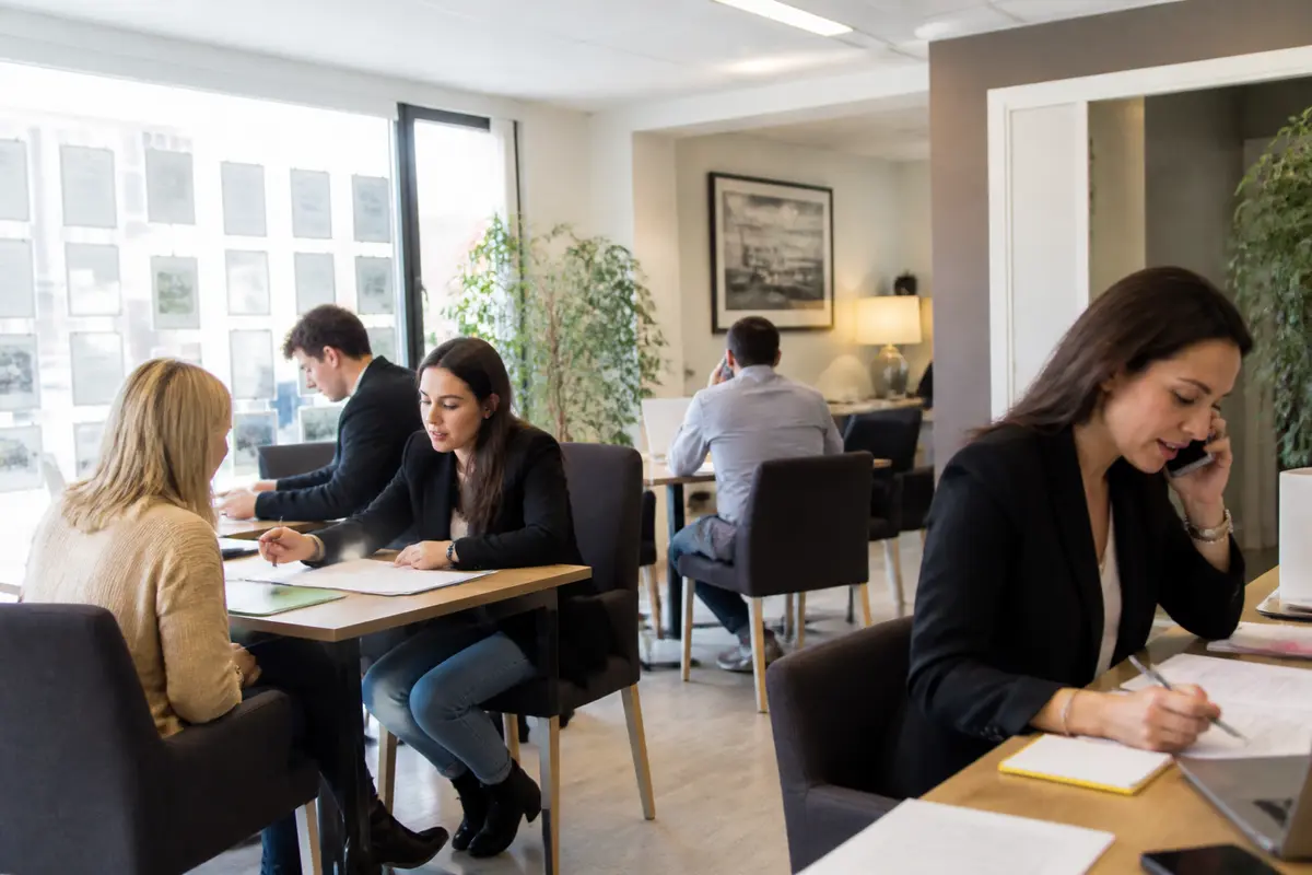 Personnes travaillant dans un bureau, discutant et utilisant des ordinateurs et téléphones. Ambiance professionnelle et collaborative.