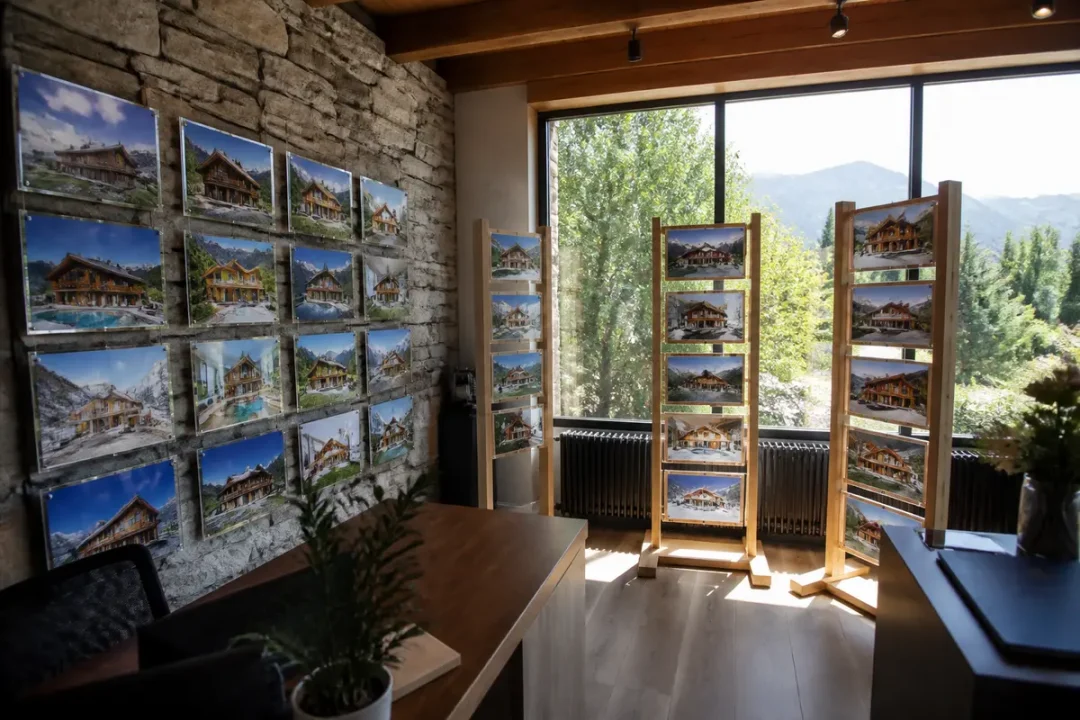 Salle d'exposition avec photos de chalets sur murs et présentoirs en bois, vue montagne, grande fenêtre.