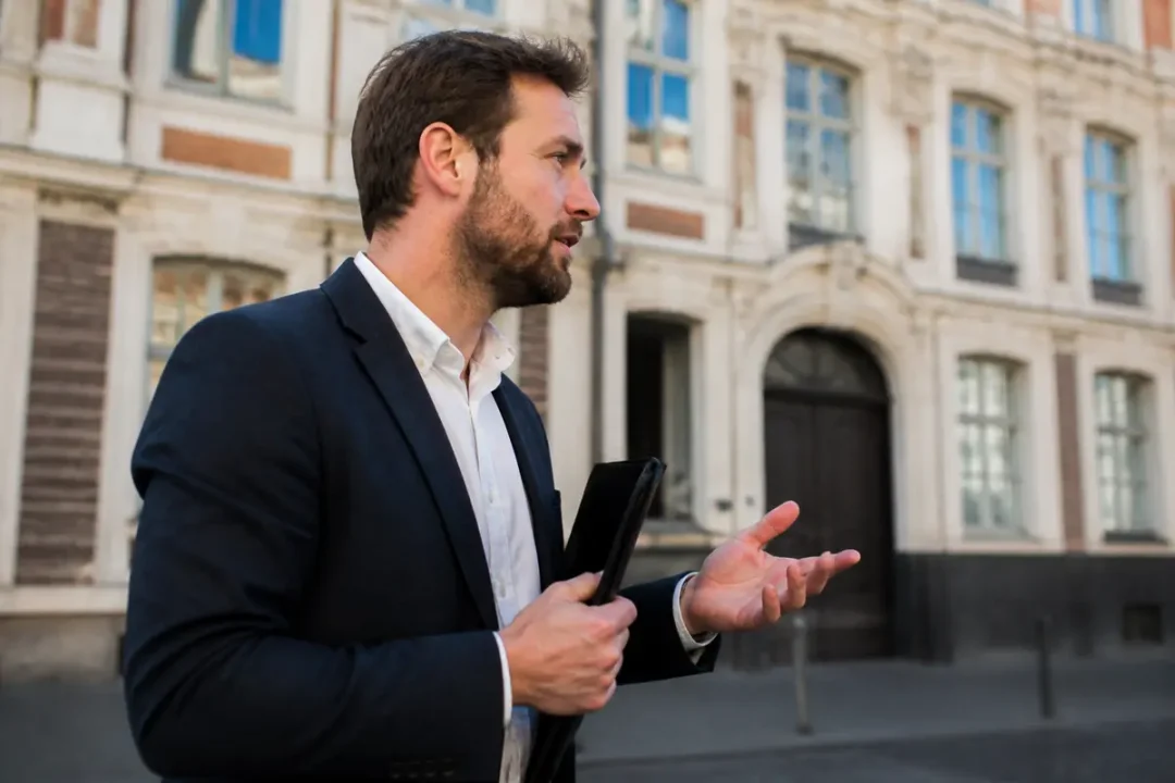 Un homme en costume parle gesticulant devant un bâtiment ancien avec fenêtres et corniches.