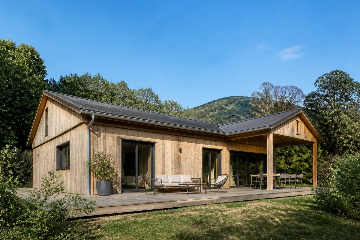 Maison en bois moderne avec terrasse, située dans un cadre naturel verdoyant, sous un ciel bleu clair.