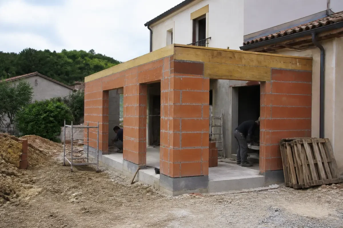 Extension de maison en construction, murs en briques rouges, échafaudage, deux ouvriers présents sur le chantier.